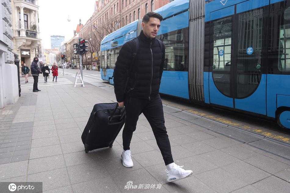 科瓦契奇手提行李，望向镜头方向，嘴角带着一丝若有若无的微笑，身旁的车流和建筑构成了背景，衬托出球星抵达时的从容与自信。