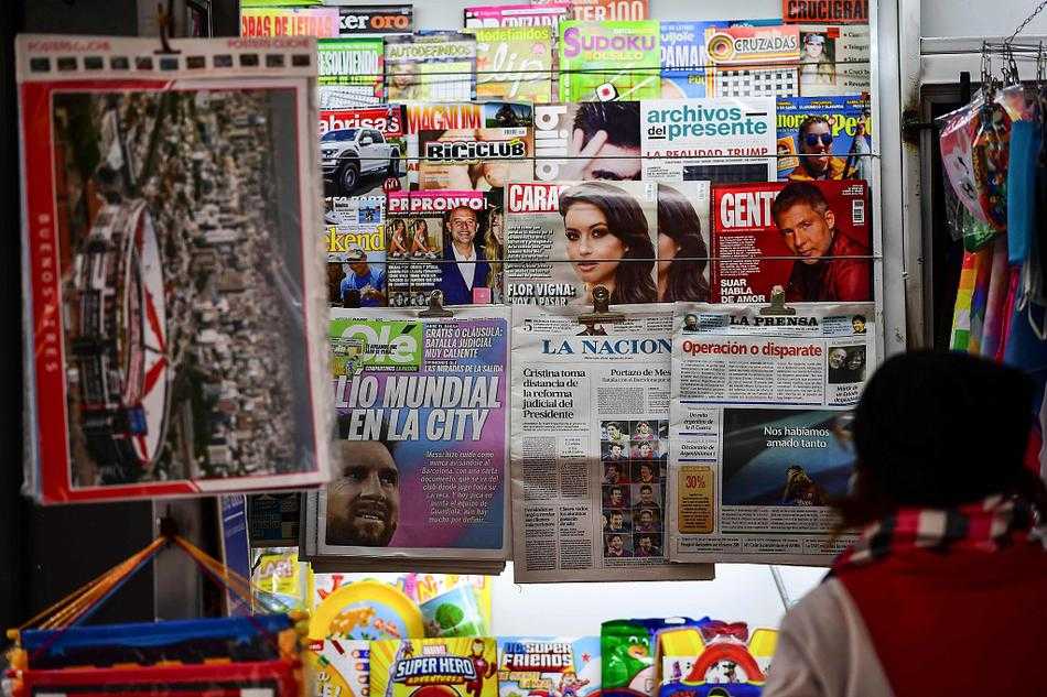 一位阿根廷读者在街边聚精会神地阅读着手中的报纸，报纸内页用整版篇幅回顾了梅西在巴萨的辉煌生涯，照片中他身穿红蓝战袍高举双臂庆祝，背景则是模糊的诺坎普球场，仿佛在诉说一个时代即将落幕。