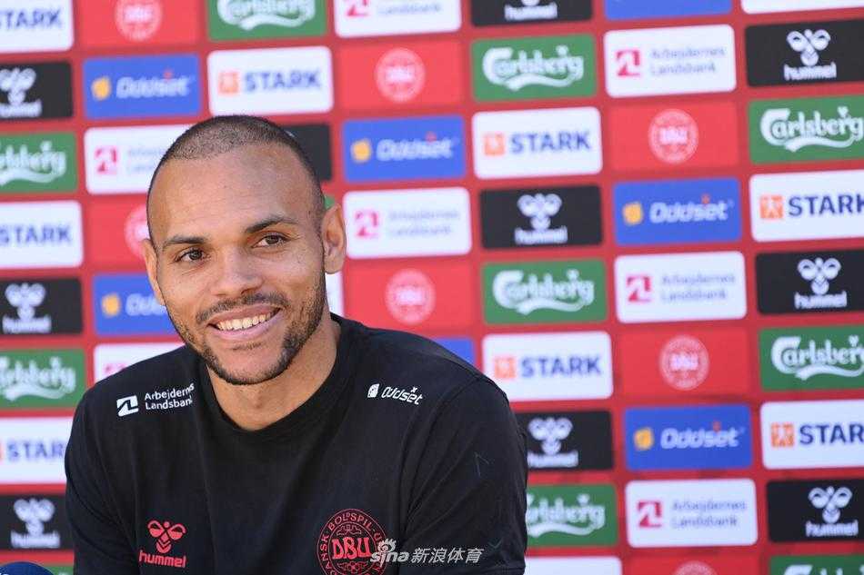 丹麦主帅尤尔曼德在新闻发布会上开怀大笑，眼角的皱纹里藏着释然与骄傲，身后的背景板印着丹麦国旗，他的肢体语言充满自信，仿佛在向世界宣告这支球队已经走出了阴霾。