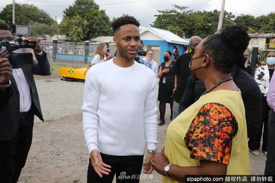 凯瑟琳公爵夫人面带微笑，站在球场边的铁丝网旁，与兴奋的当地民众亲切握手互动，她鲜艳的条纹连衣裙在阳光下格外亮眼。