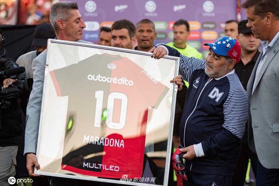 身披深色大衣的马拉多纳，面带标志性的微笑，在客队球迷看台前高举双臂鼓掌致意，仿佛一位指挥家，引领着全场山呼海啸般的欢呼与掌声。