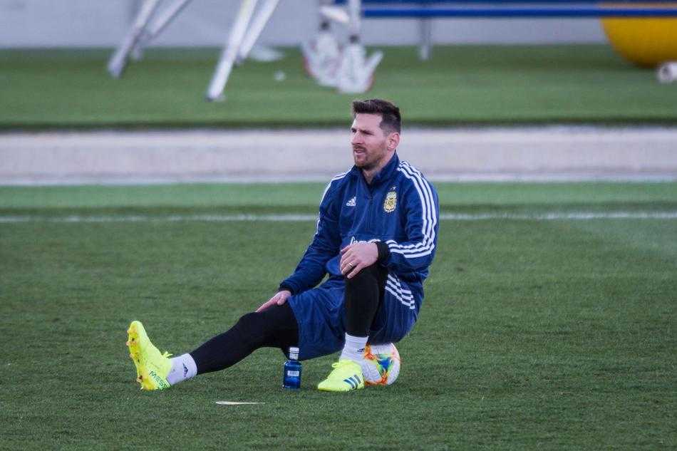 训练间歇，梅西坐在草地上，用双脚灵活地夹住足球并抬起，脸上带着轻松愉快的表情，与身旁的队友德保罗谈笑风生。