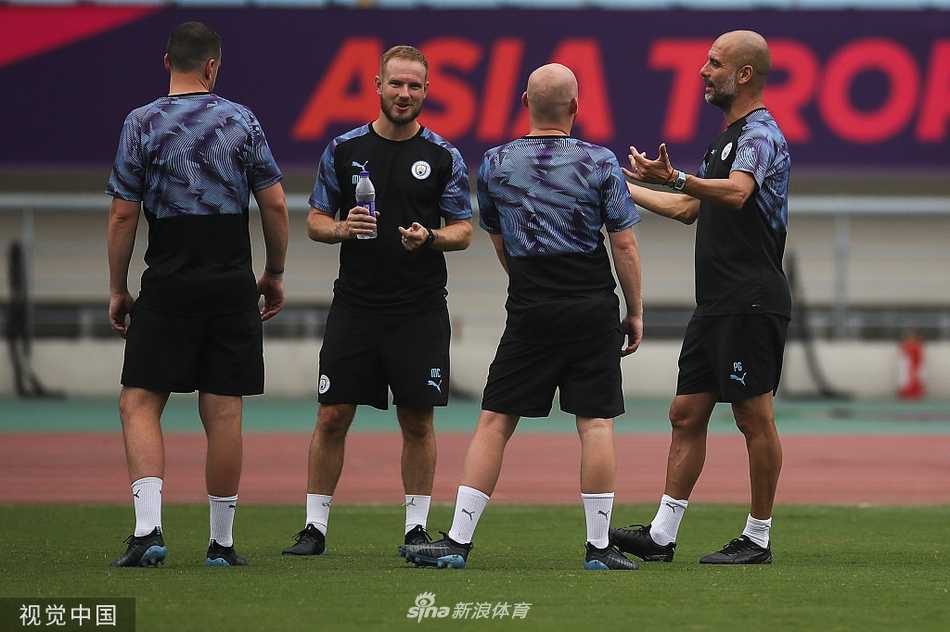 瓜迪奥拉在南京训练场上专注指挥，双手摊开，仿佛正对某个未达预期的跑位提出质疑，身后是湛蓝的天空和训练场设施。