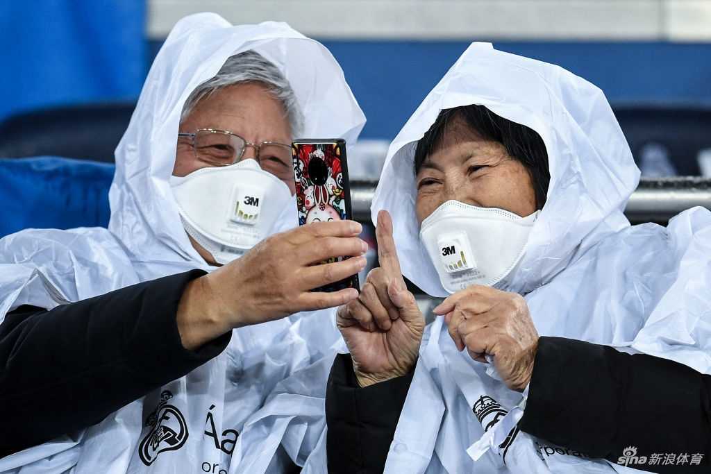 看台上，一名戴眼镜的亚裔男青年紧裹着深色外套，白色的口罩遮住了大半张脸，他双手轻轻放在膝盖上，仿佛在喧闹的球场中守护着自己的独立小世界。