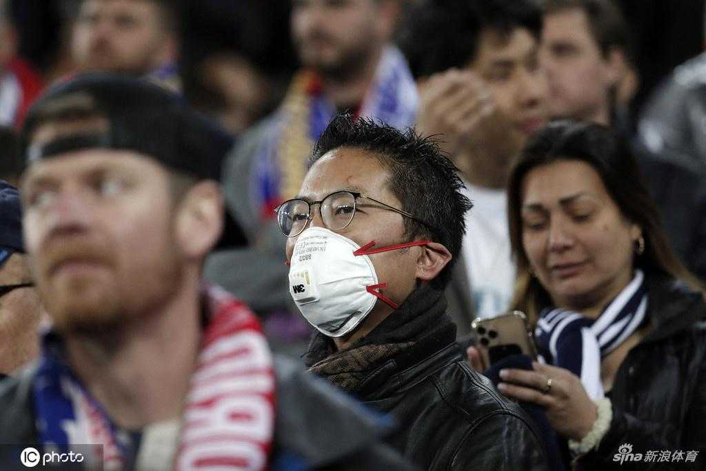 一位亚裔女球迷在沸腾的伯纳乌看台上，用口罩将自己与喧嚣暂时隔离，眼神专注地望向球场方向，蓝白色的织物与身后的白色海洋形成鲜明对比。