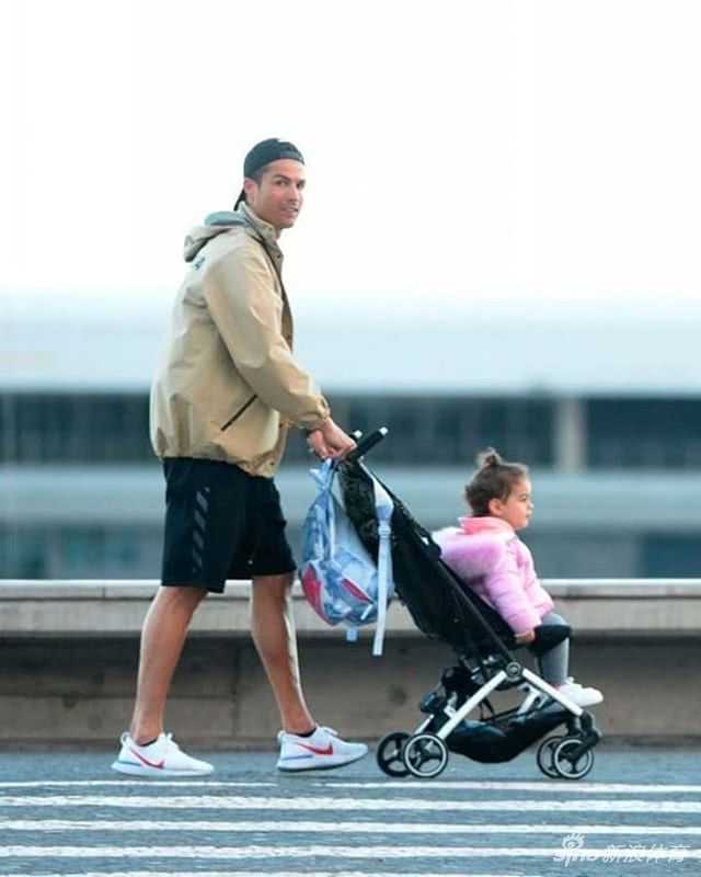 C罗戴着黑色棒球帽，身穿简单的深色运动装，正低头温柔地注视着婴儿车里的孩子，一旁的乔治娜也俯身整理衣物，两人全身心都扑在孩子身上，完全没注意到远处的镜头。