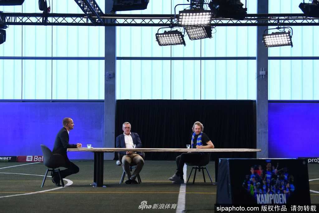 几名布鲁日球员戴着口罩，保持着社交距离，围在一起看着手机屏幕上队友捧杯的画面，虽然面容被遮挡，但眼神中流露出的兴奋与笑意清晰可见。