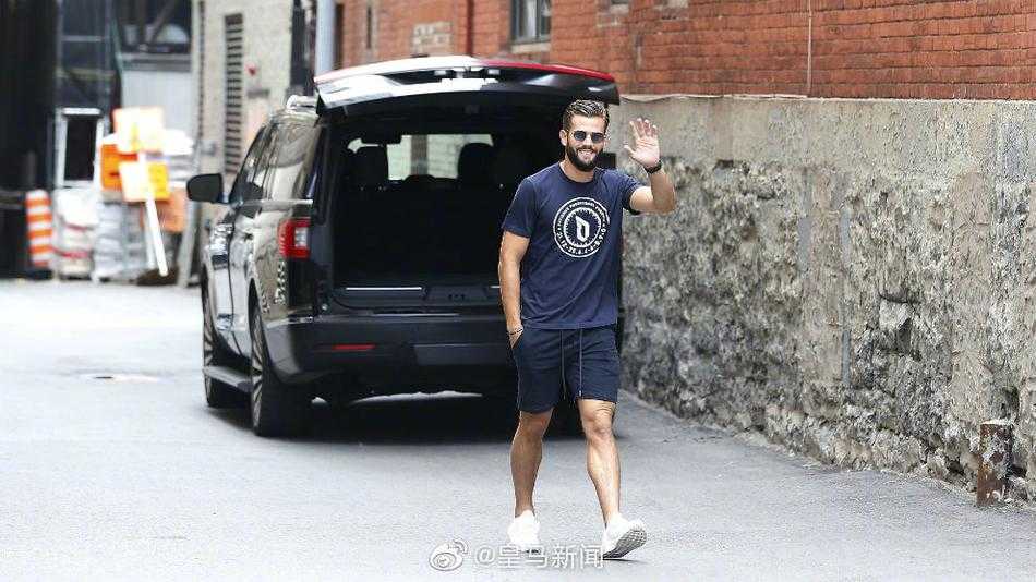 镜头拉近，久保建英身穿深色休闲上衣，眼神清澈看向前方，一旁的卢宁戴着墨镜，两人正穿过一个带有拱门的街区，背景是充满西班牙风情的建筑。