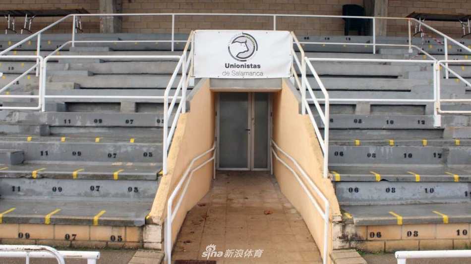 从球场草坪一角望去，远处的单层主看台在空旷的场地中显得格外醒目，跑道上的白色线条清晰可见，整个场景充满了朴素而真实的足球氛围。