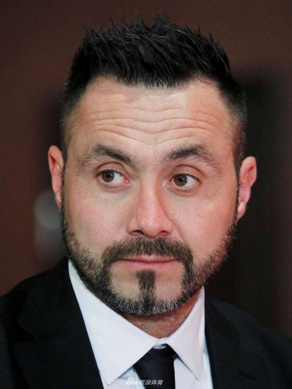 德泽尔比手持顿涅茨克矿工的蓝色围巾，面带微笑望向一侧，仿佛在向热情的矿工球迷致意。