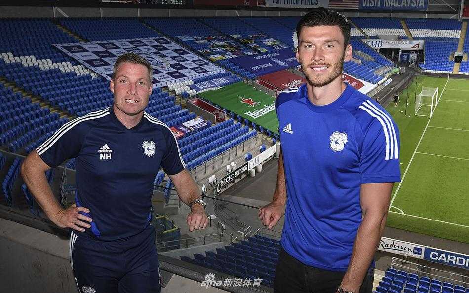 身高195厘米的威尔士高中锋基弗·穆尔在签约仪式上，双手持卡迪夫城蓝色主场战袍，面对镜头展露自信笑容，背景是简洁明亮的摄影棚。