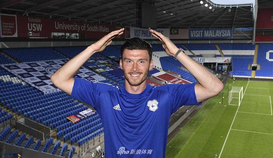 在卡迪夫城训练场的草坪上，基弗·穆尔完成签约后与主教练尼尔·哈里斯并肩站立，两人一同展示签有穆尔名字的28号球衣，背景是空旷的看台，预示着即将在这里为荣誉而战。