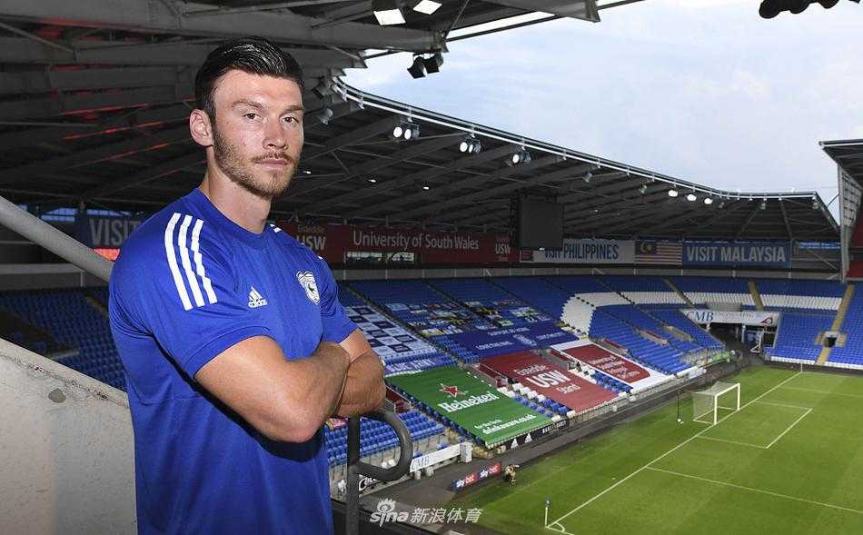 基弗·穆尔坐在卡迪夫城俱乐部内的复古看台座椅上，轻松地秀着新赛季的蓝色战袍，他身姿舒展，背景是看台通道和金属栏杆，画面充满生活气息和即将开启新篇章的期待感。