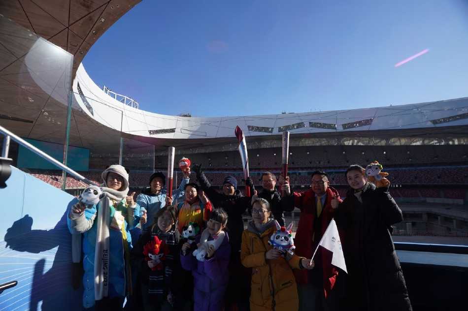 冰立方冰上运动中心内，可爱的吉祥物“冰墩墩”人偶被一群活泼的小朋友团团围住，孩子们伸出小手试图与它拥抱，冰墩墩则憨态可掬地弯下腰，与孩子们进行温馨互动，画面充满童真与喜悦。