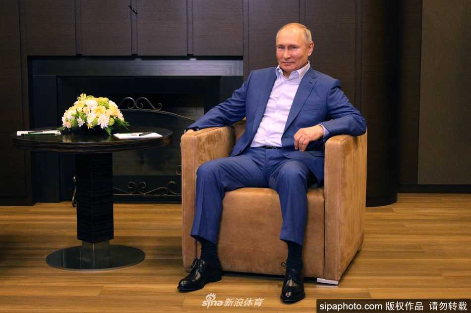 镜头拉近，捕捉到了交谈中的生动细节。法塞尔主席身体微微前倾，双手交叠，面带专注的笑容，似乎正在回应普京关于场馆建设的议题。桌上摆放的物品和窗外的绿意，为这场严肃的会谈增添了一丝生活气息。