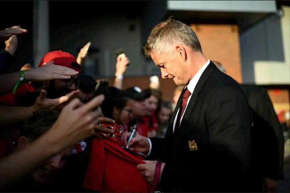 索尔斯克亚面带微笑，与身边的曼联球员们一起在球迷区与热情的支持者们亲密合影，背景是老特拉福德球场标志性的看台，现场气氛热烈而融洽