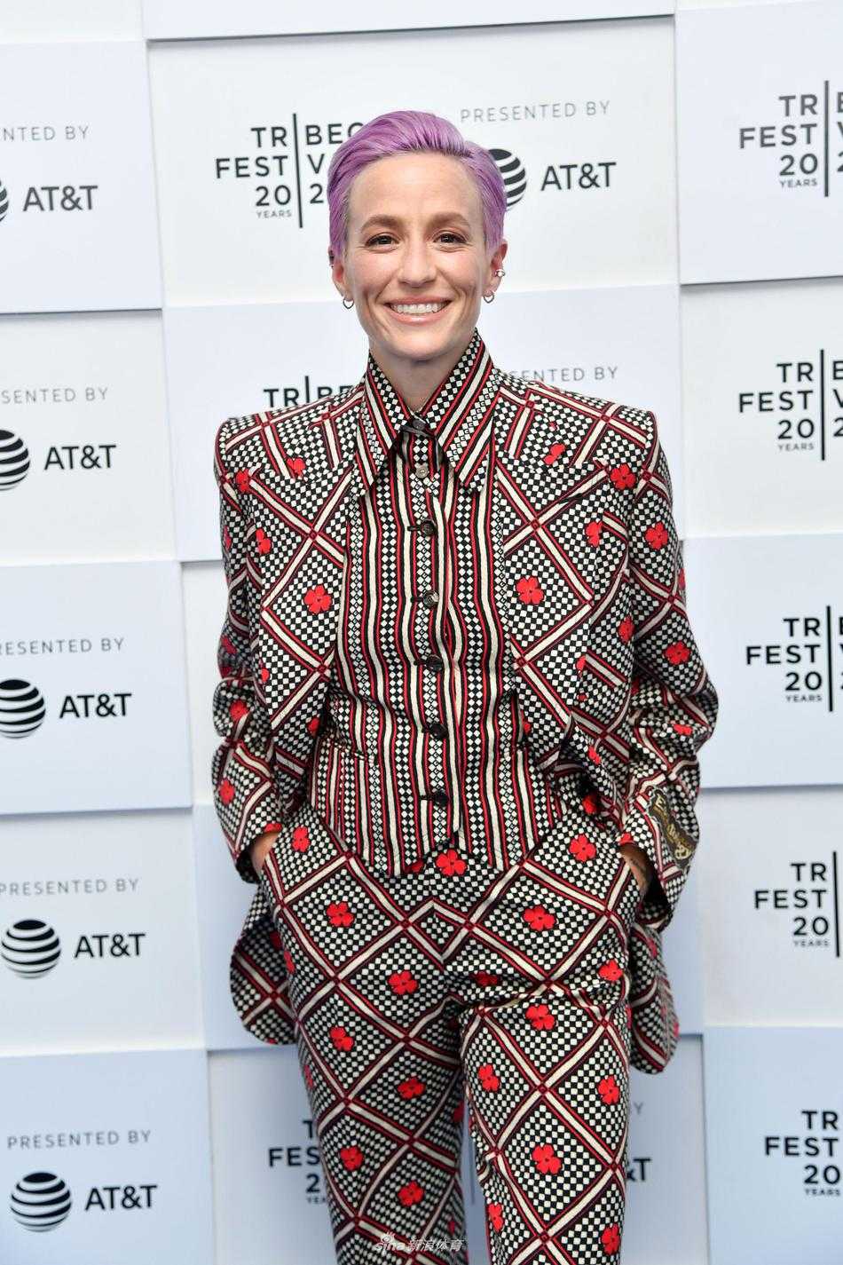 梅根·拉皮诺埃身着一套色彩斑斓、剪裁利落的花色西装，自信地站在翠贝卡电影节的背景板前，她那标志性的粉紫色短发与身上的色彩相映成趣，仿佛一幅行走的艺术品。