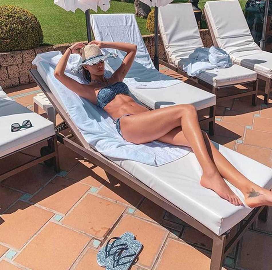 乔安娜·桑兹身着深色连衣裙，倚靠在墙边，深邃的眼眸凝视镜头，举手投足间散发着慵懒而自信的迷人气质。